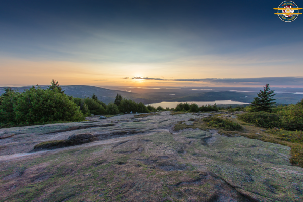 Sunrise from Cadillac Mountain – Acadia National Park Sunrise view from a rocky mountain summit overlooking forested hills, lakes, and distant coastline.