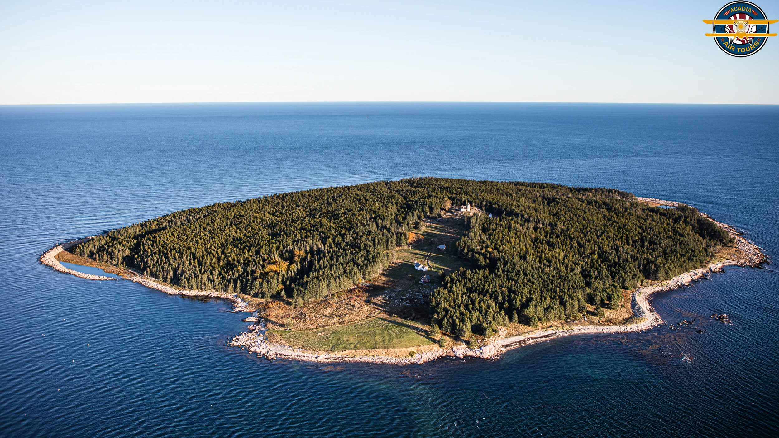 Aerial view of a forested island surrounded by deep blue ocean, with clear shoreline and scattered small buildings.