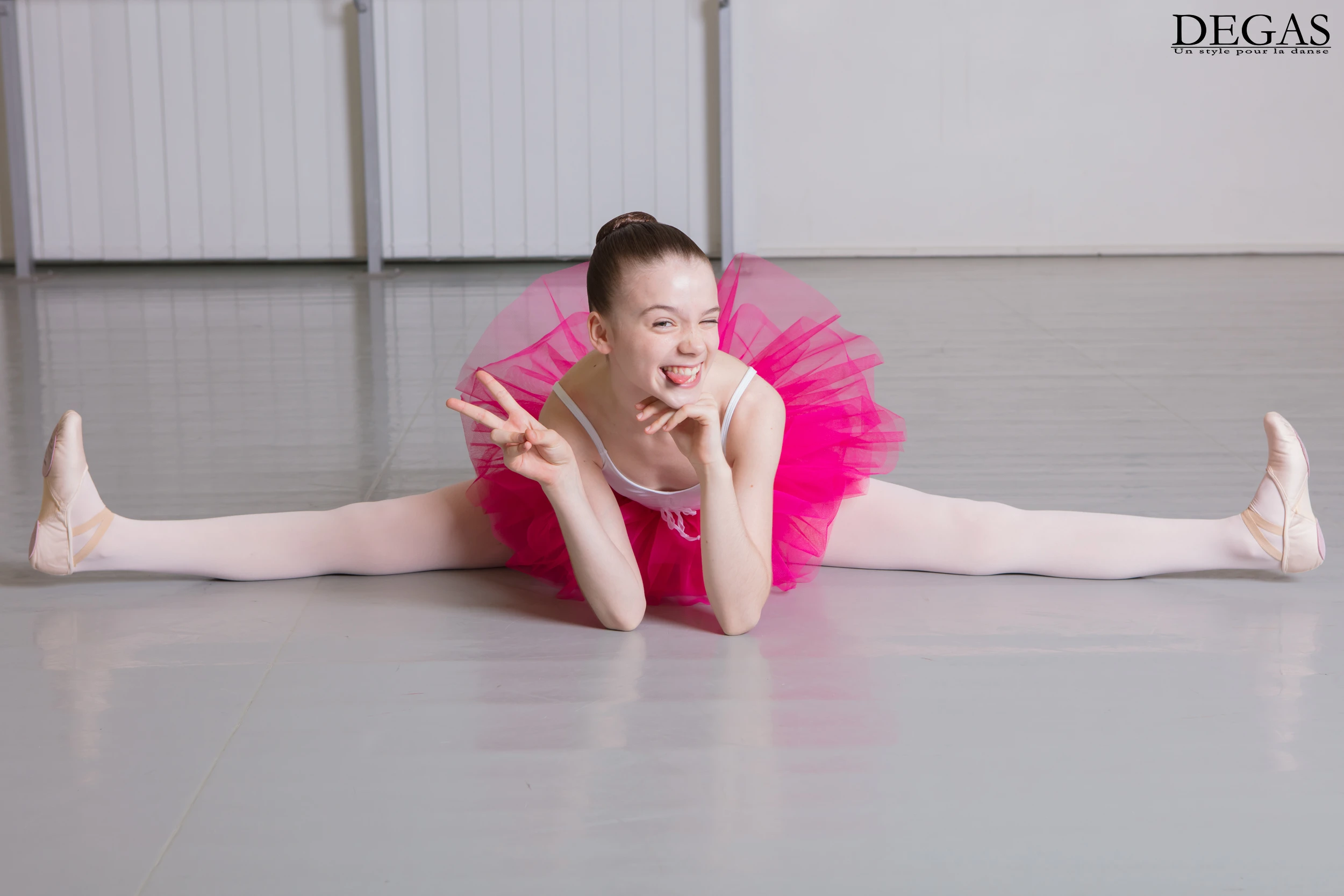 Young ballerina in pink tutu doing a full middle split while smiling and winking at the camera.