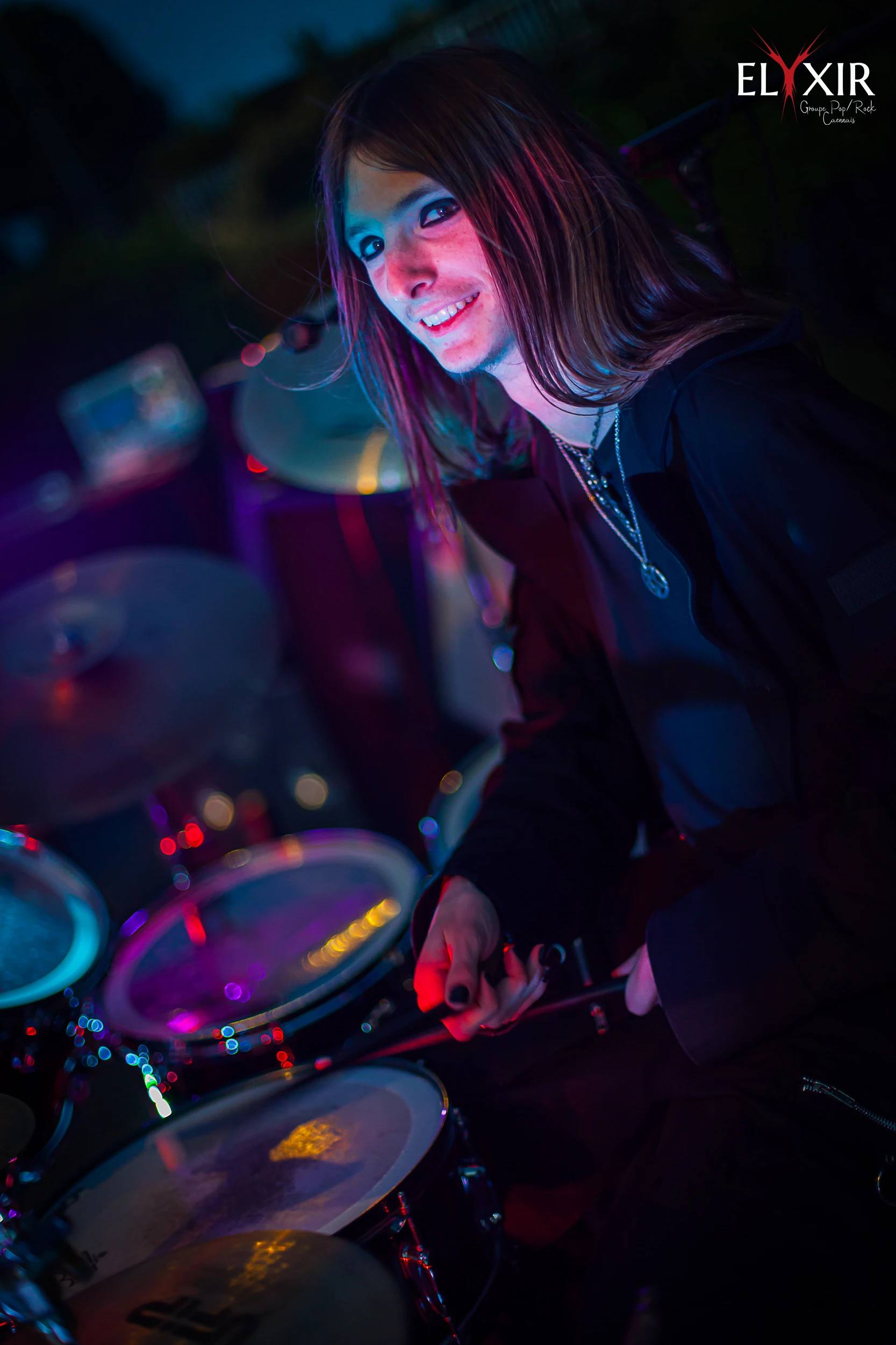 Drummer smiling during a live concert, surrounded by a vibrant and colorful drum kit under stage lighting.