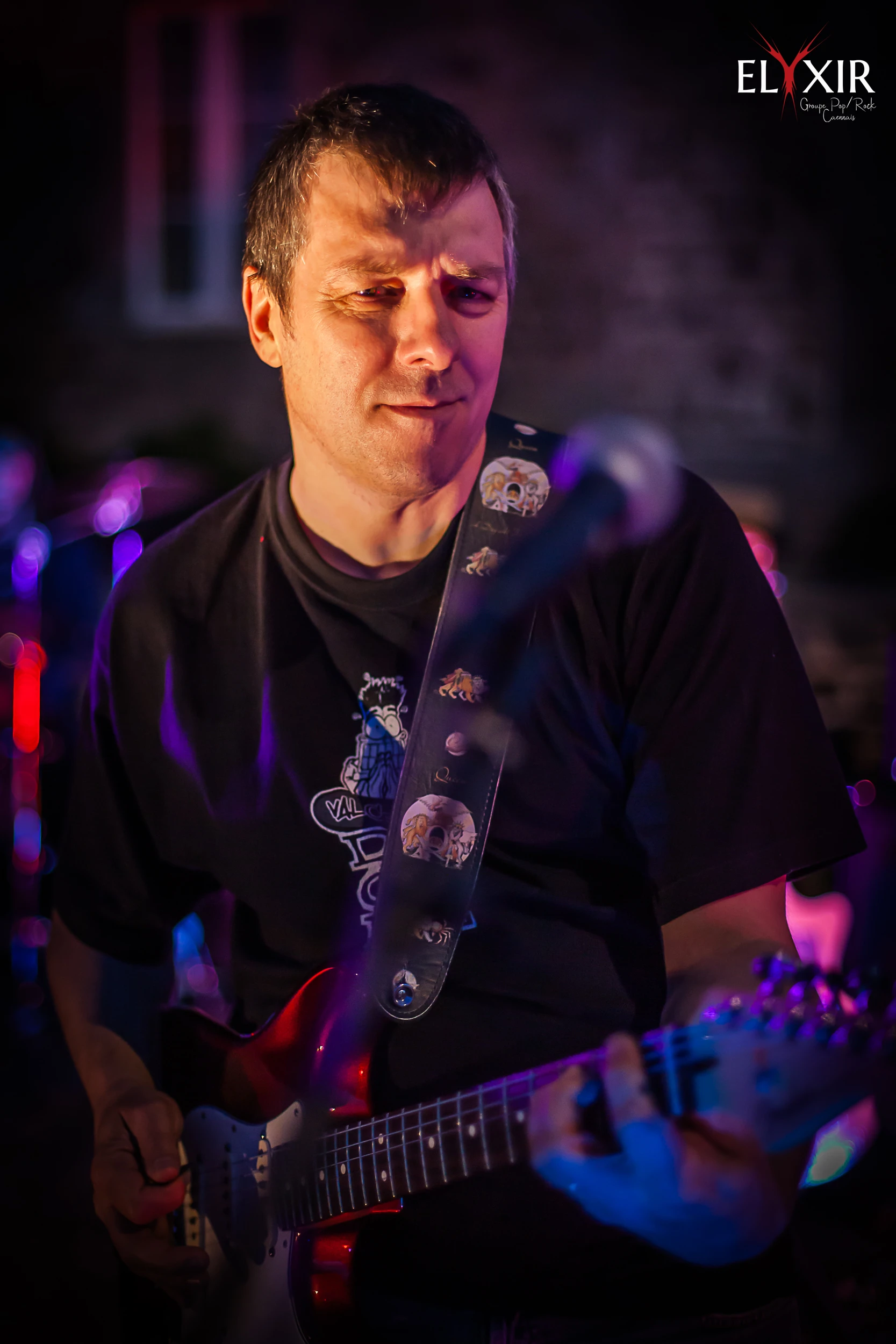 Guitarist Xav performing live on stage, illuminated by colorful stage lighting, with expressive focus on his electric guitar.