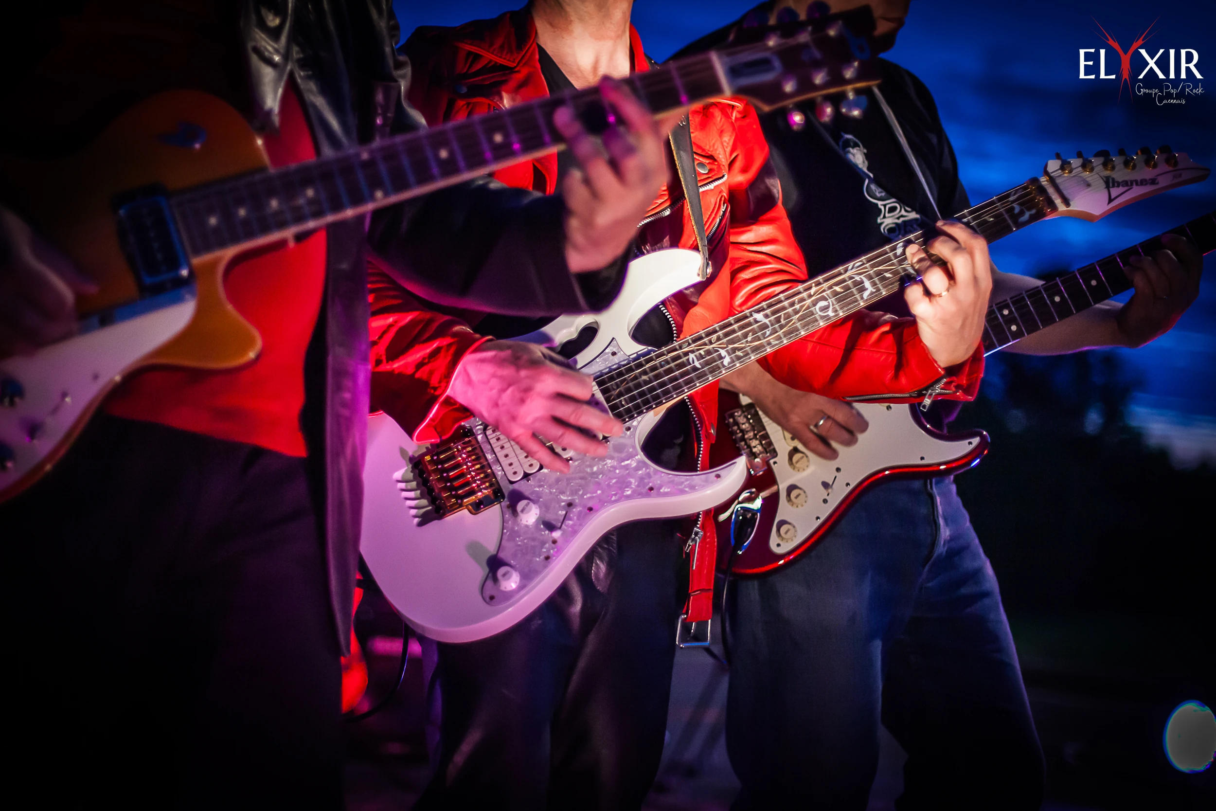 Close-up of three guitarists playing electric guitars under concert lighting during a live performance.