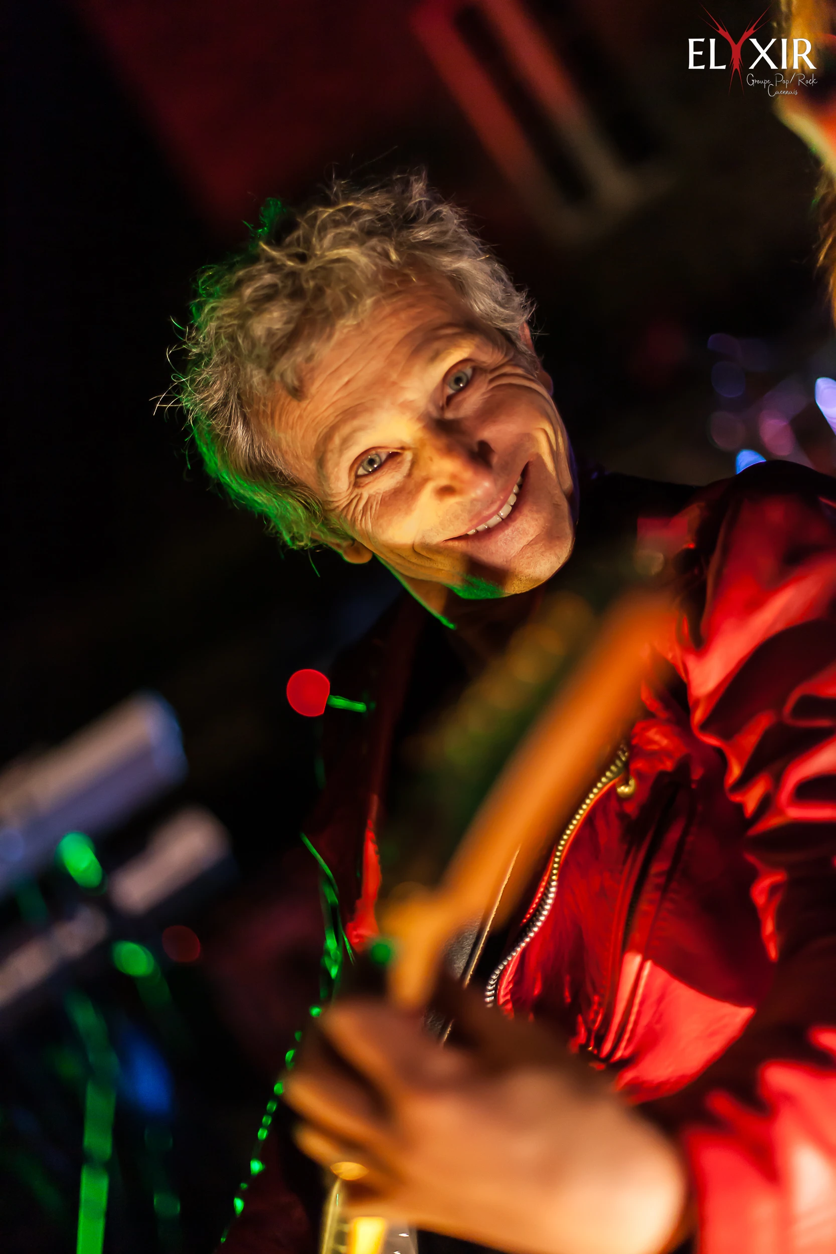 Philip, guitarist of the band Elyxir, smiling toward the camera under colorful concert lighting during a live performance.