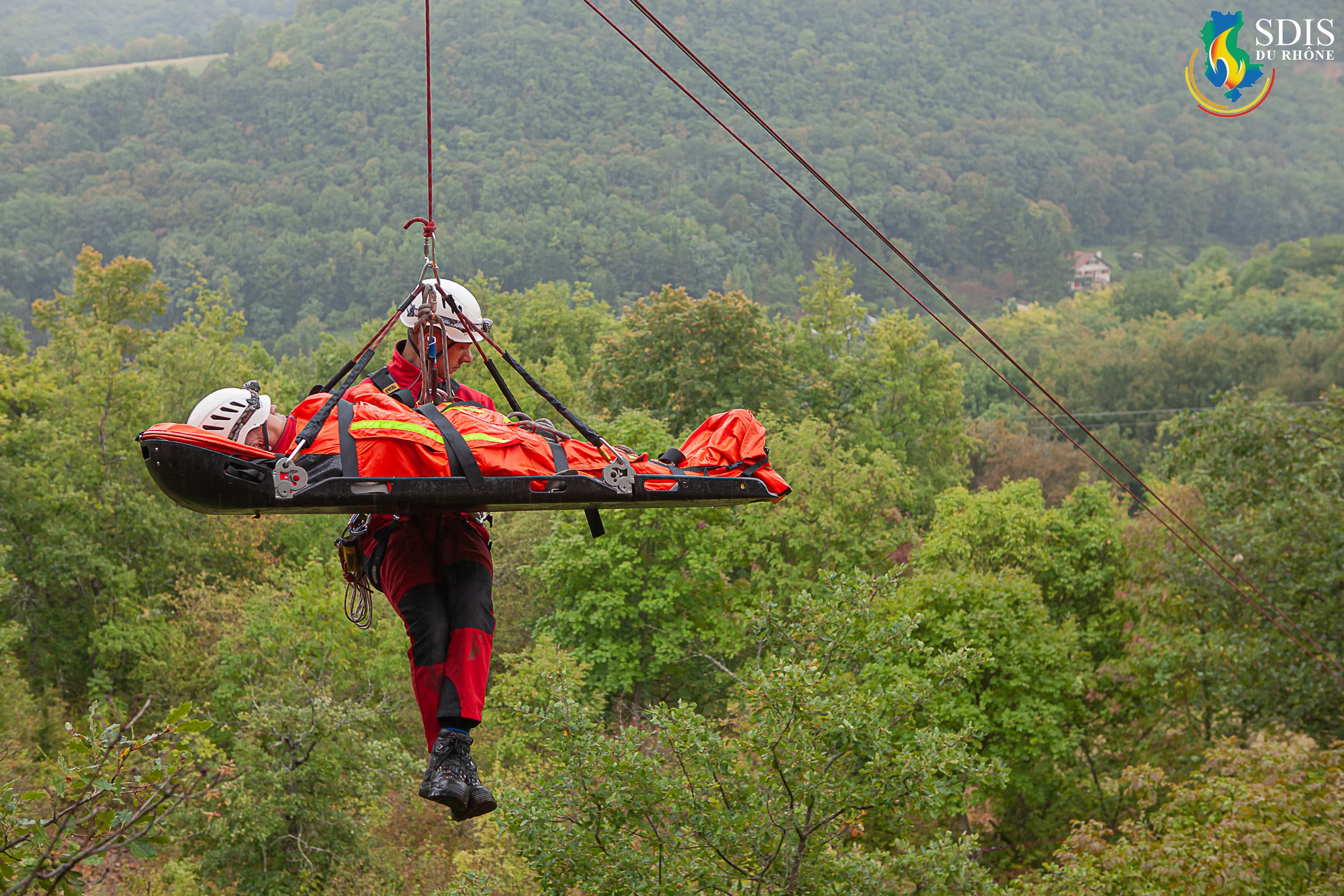 GRIMP firefighter from SDIS du Rhône performing a rescue drill on a stretcher over a forested area.