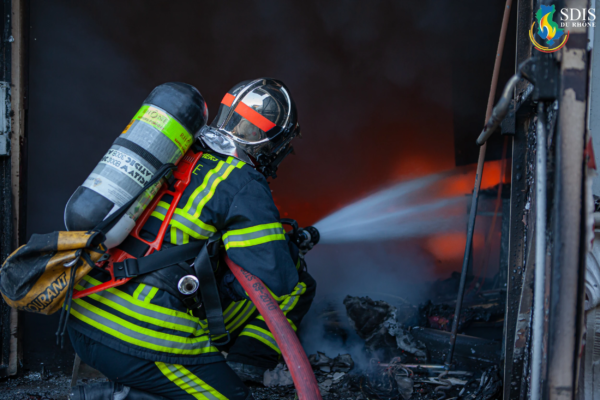 Close-range fire attack by SDIS du Rhône firefighter during warehouse blaze Firefighter from SDIS du Rhône kneeling while spraying water into a burning warehouse filled with debris.