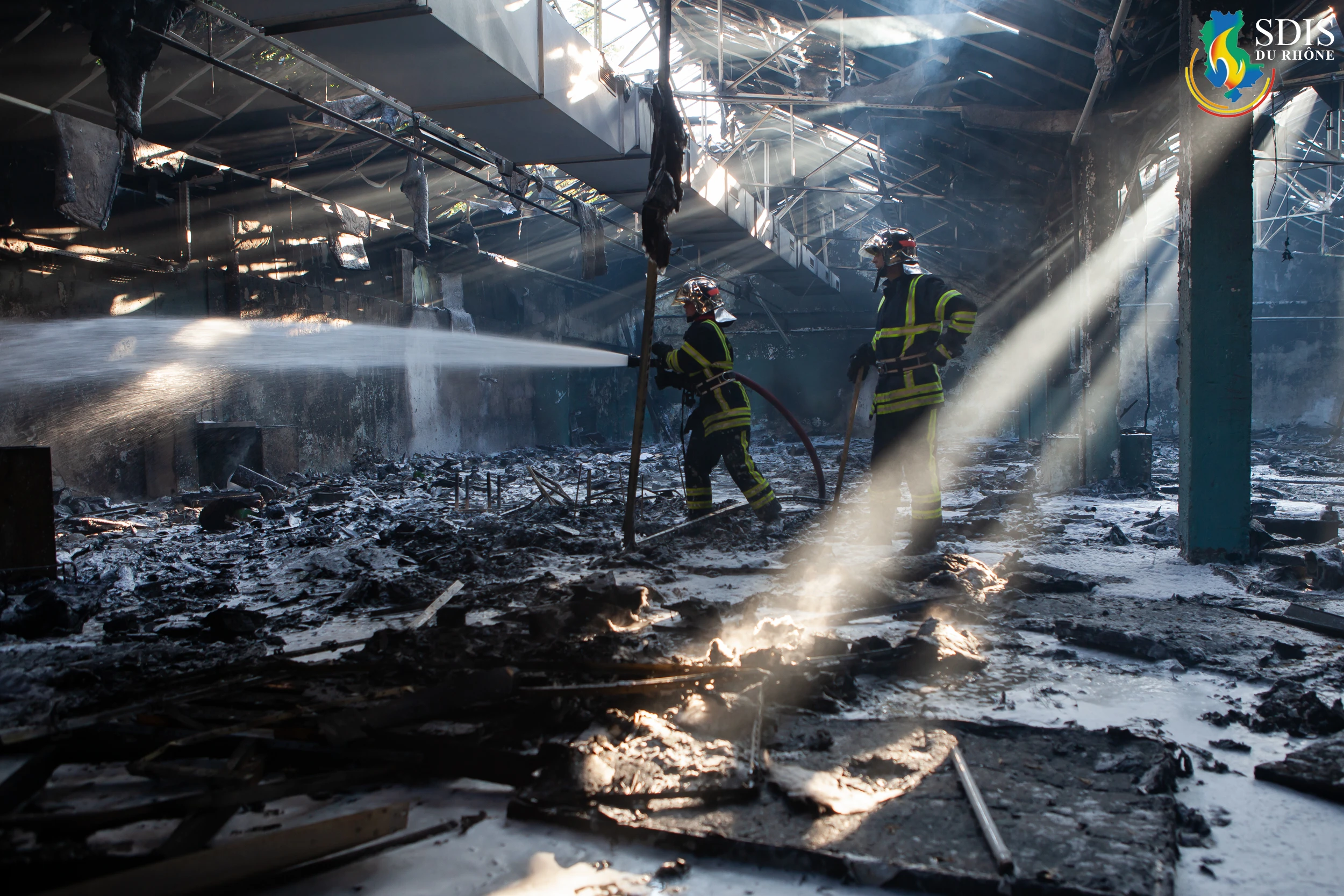 Firefighters from SDIS du Rhône extinguishing a massive warehouse fire amidst smoke and destruction.