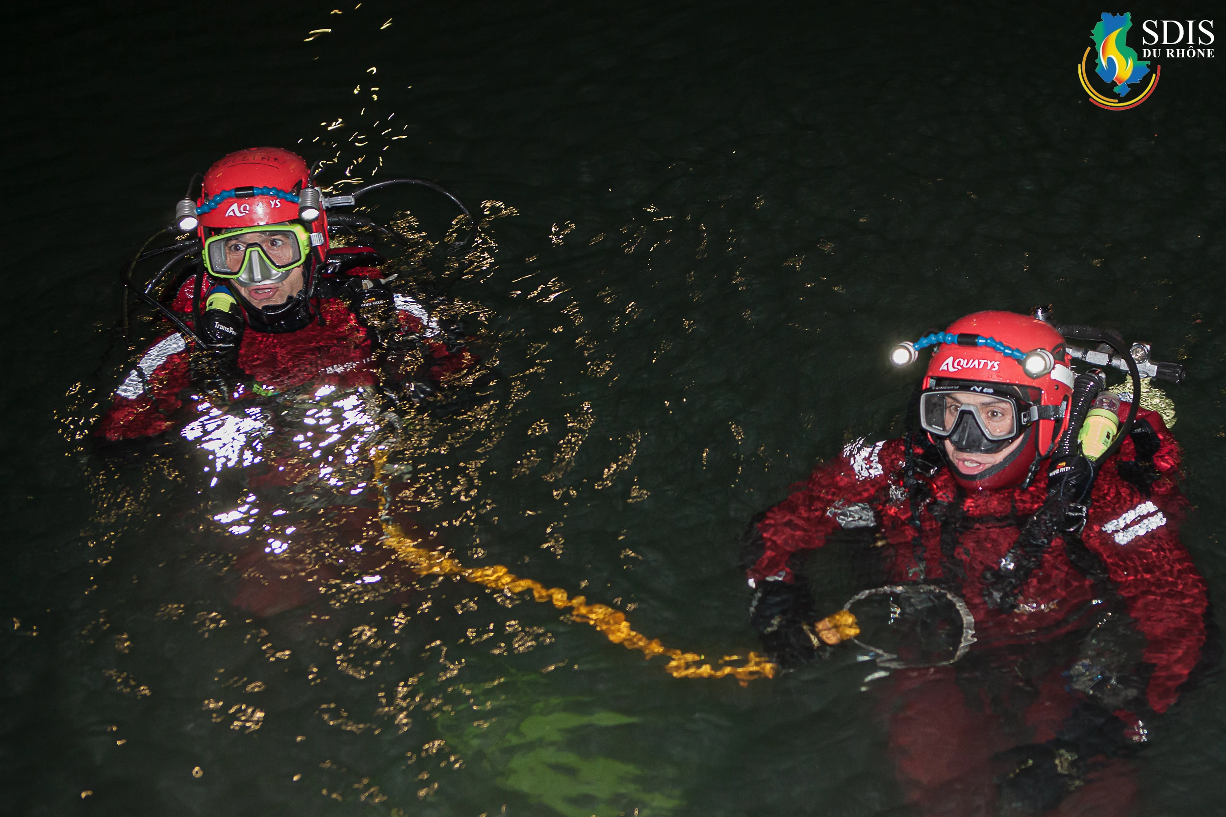 Two SDIS Rhône divers in full red rescue gear during a nighttime search operation in the Rhône River, connected by a safety line.