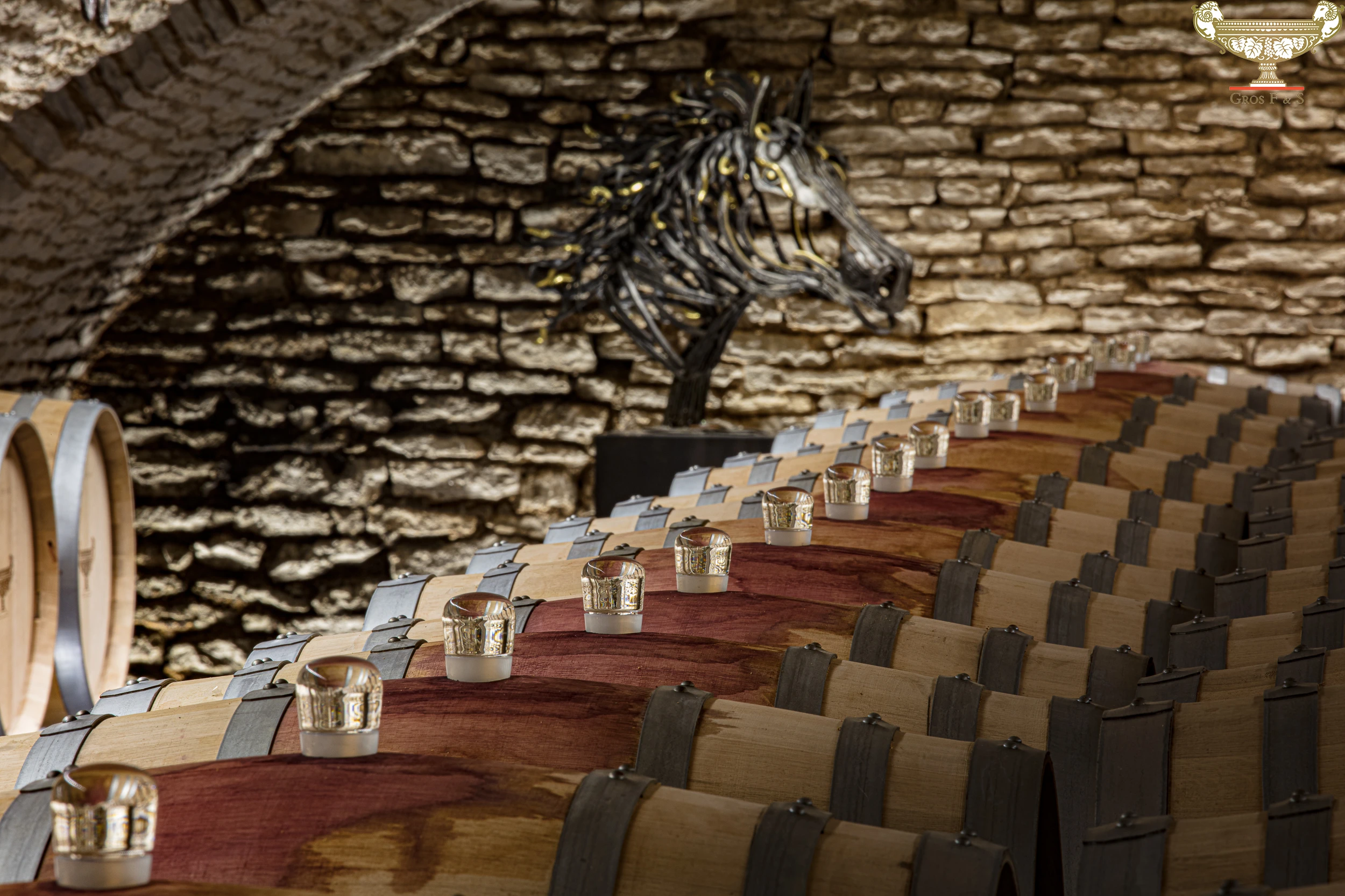 lose-up of wine barrels with glass airlocks in a vaulted cellar at Domaine Gros Frère et Sœur, with a metallic horse sculpture in the background.