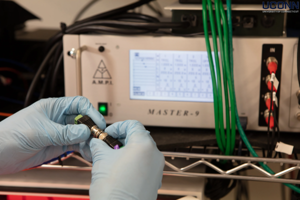 Precision Wiring for Neural Research Equipment – UConn Lab Close-up of gloved hands connecting a black coaxial cable to a piece of neuroscience research in a UConn laboratory.