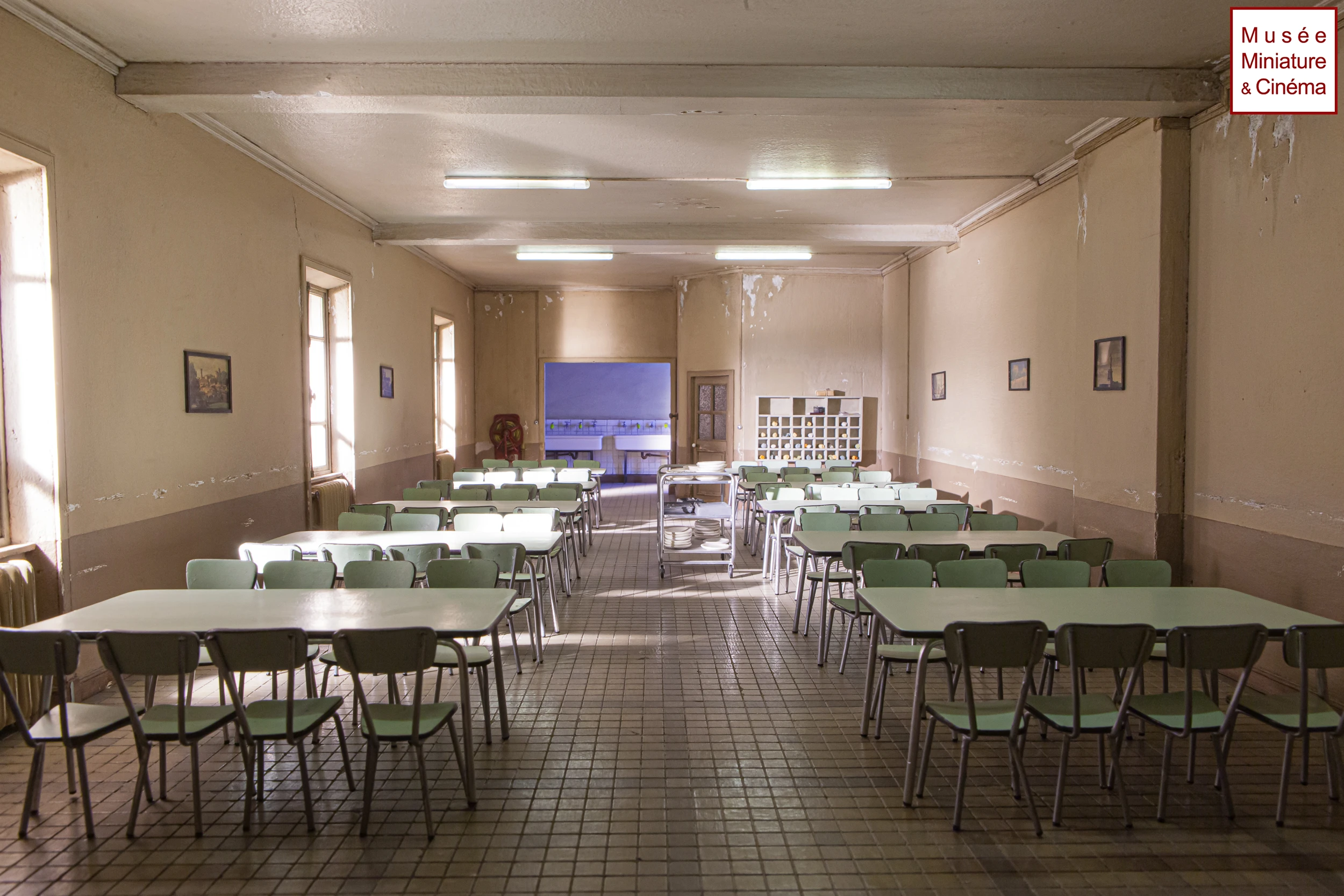 Miniature reproduction of a vintage school cafeteria with green chairs, white tables, and tiled floor.