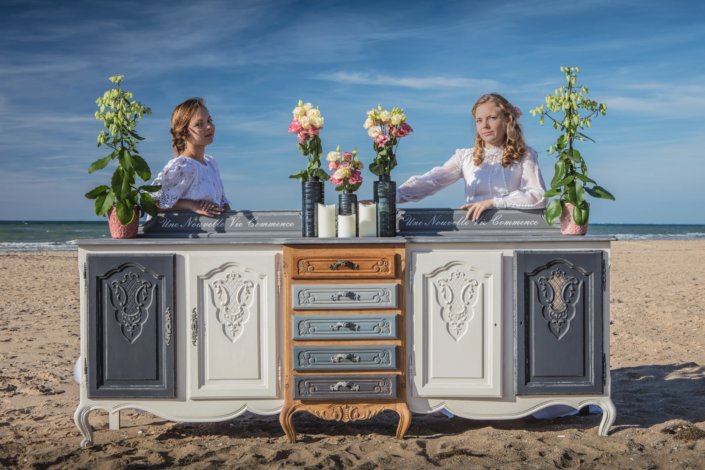 Two women in white dresses with floral decor and repainted sideboard on the beach — creative visual identity by Atelier Gris Mousse.