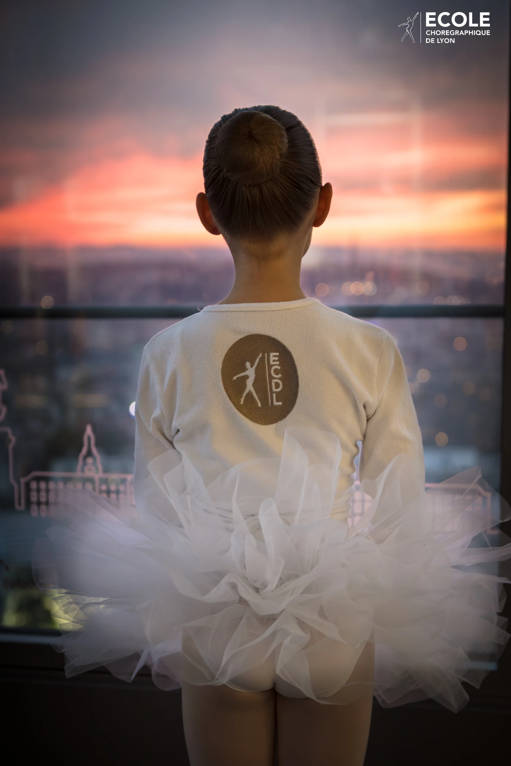 Young ballet dancer in ECDL attire facing the Lyon skyline at sunset.