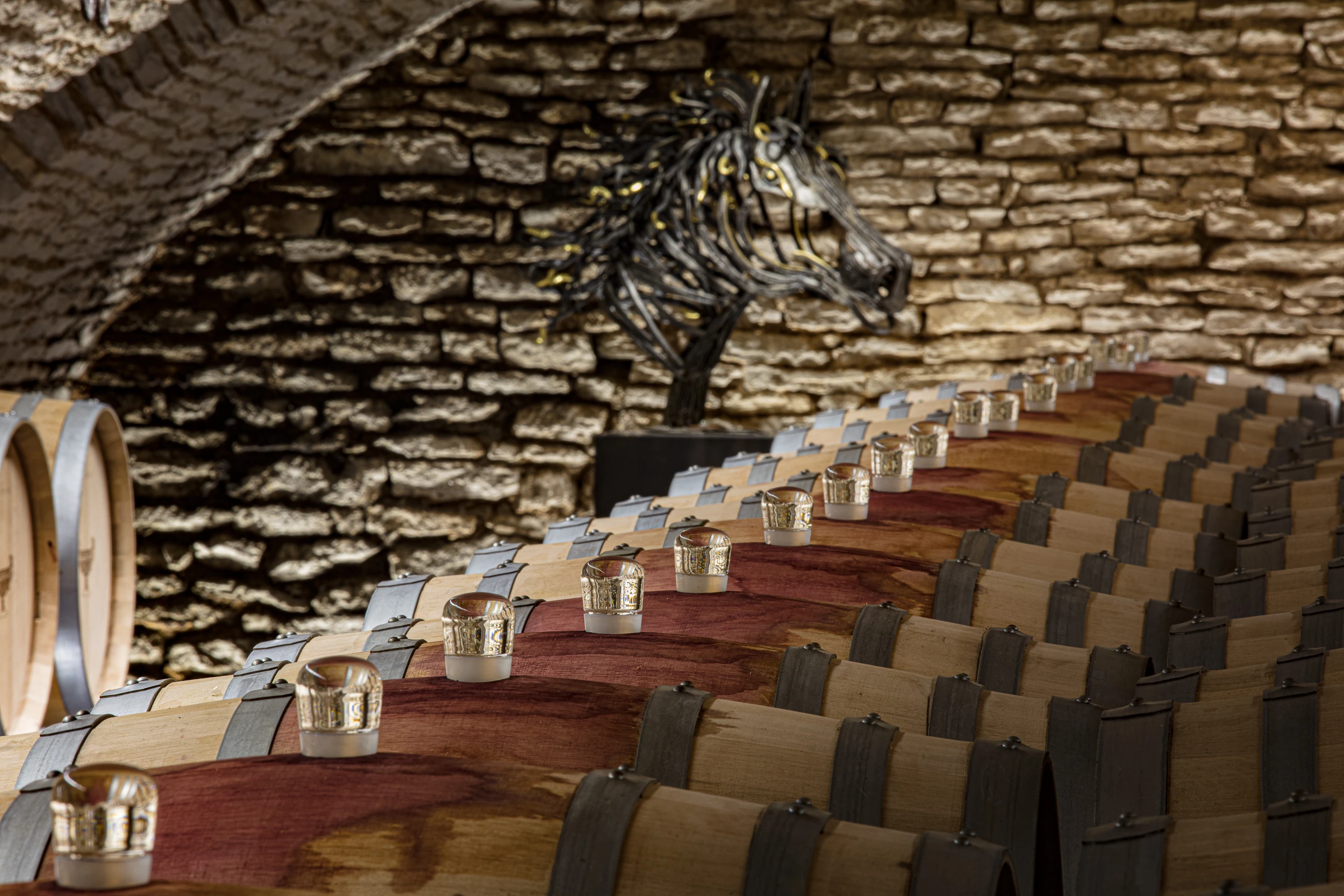 lose-up of wine barrels with glass airlocks in a vaulted cellar at Domaine Gros Frère et Sœur, with a metallic horse sculpture in the background.