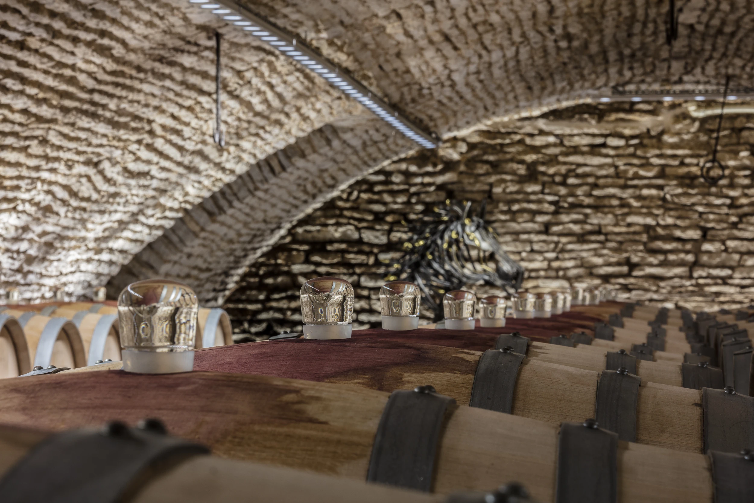 Oak barrels lined up under stone arches in the cellar of Domaine Gros Frère et Sœur, with a metal horse sculpture in the background.