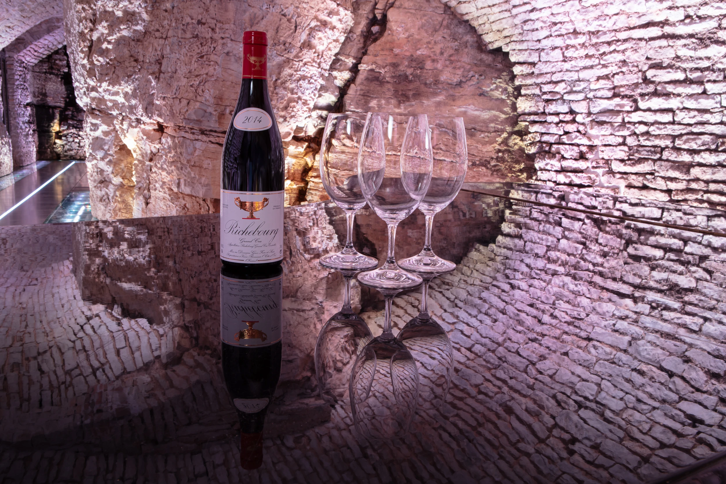 Bottle of 2014 Richebourg Grand Cru wine with four empty glasses on reflective glass table in a historic Burgundy wine cellar.