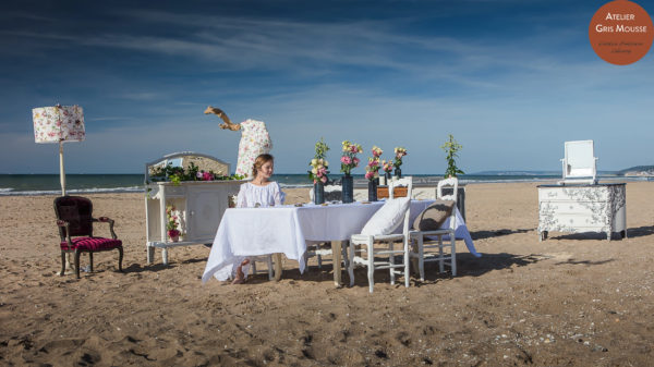 Restyled vintage dining set staged on the beach at golden hour, a young woman in a white lace dress seated at the table