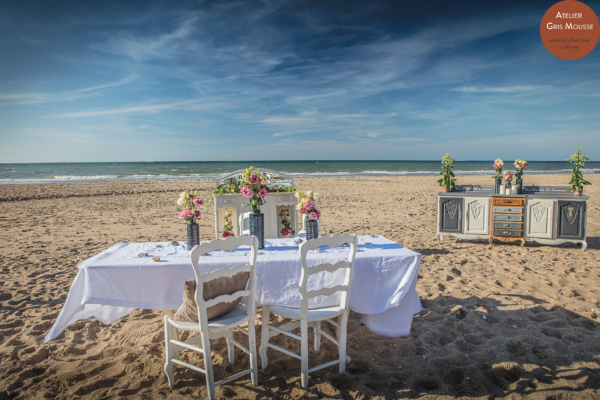 White-clothed dining table with two restyled chairs facing the sea, a painted sideboard behind
