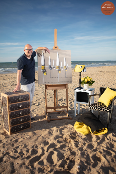 Painter Christian Bellec standing beside his canvas on an easel on the beach, next to a restyled anchor-patterned chest and a black and yellow styled corner
