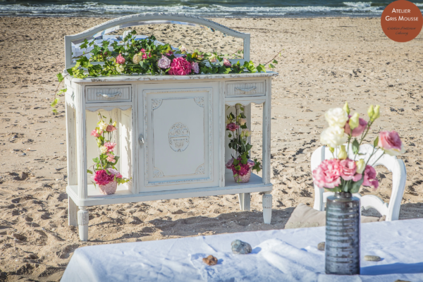 White patinated marble-top sideboard dressed with ivy and roses, standing on the sand by the sea.