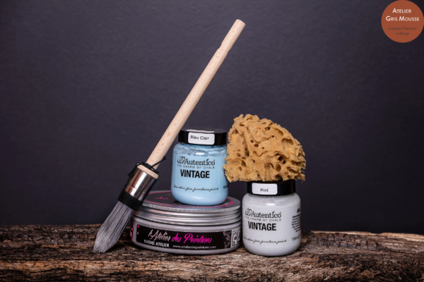 Autentico Vintage chalk paint jars in Bleu Clair and Mint, an Atelier des Peintures patina tin, a round brush and a natural sponge on a log