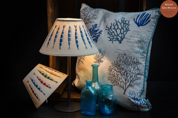 Lamp with a sardine-print shade beside a coral-print cushion and translucent blue glass vases, coastal boutique styling