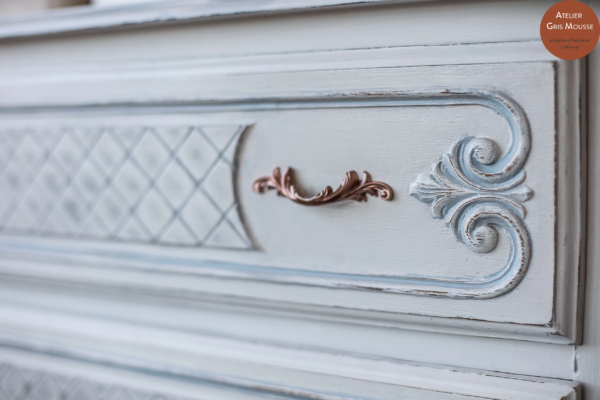 Close-up of a pale blue-grey patinated drawer with a rose-gold handle, carved scrolls and a lattice panel