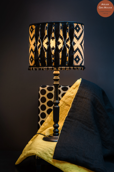 Black table lamp with a backlit geometric handmade shade, a polka-dot chair and yellow and black throw behind