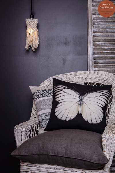 White rattan chair with a black butterfly-print cushion and graphic cushions, a macramé pendant light beside it