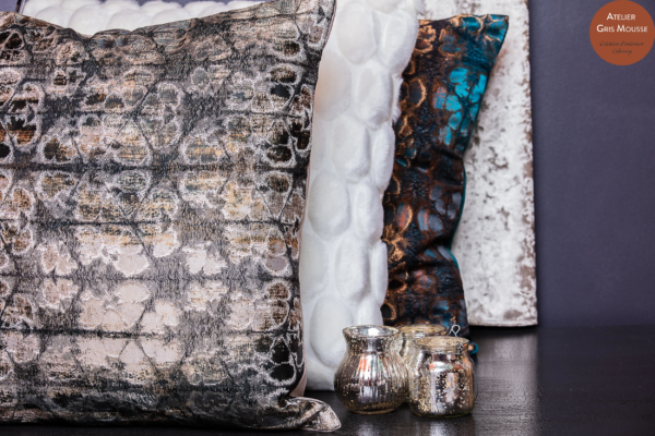 Stack of textured cushions in silver, gold, white faux fur and teal velvet beside small silver-painted glass candle holders
