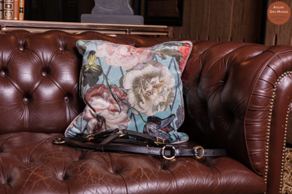 Floral velvet cushion on a brown leather Chesterfield with a leather halter laid across the seat, bookshelf behind