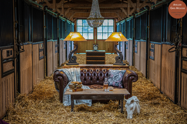 Leather Chesterfield sofa, crystal chandelier and lamps set up along the aisle of a working stable, a horse-head sculpture on the straw
