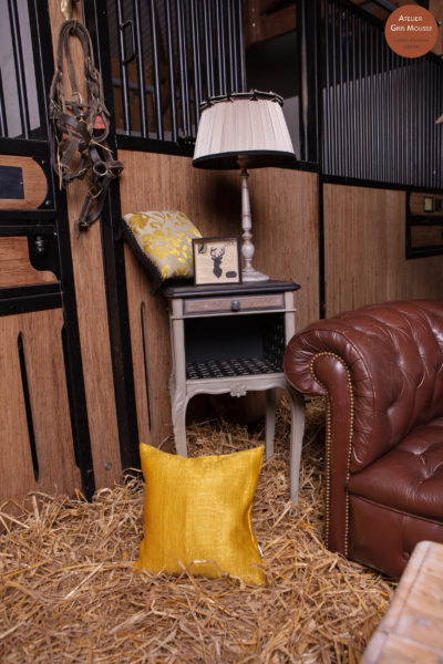 Restyled grey nightstand with a stag-motif lamp and framed print, a yellow cushion and Chesterfield in a stable corner
