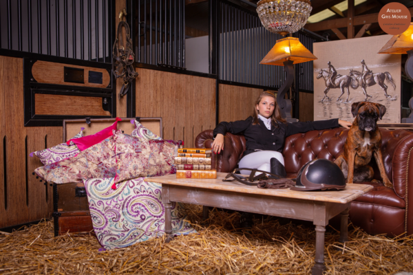 Young rider in competition dress seated on a leather Chesterfield in a stable, a boxer dog beside her and equestrian decor around